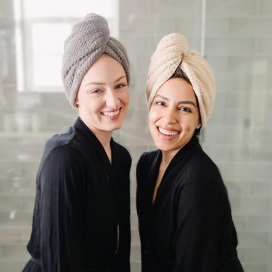 Two smiling women wearing hair towel wraps twisted on their heads, one in gray and one in beige, both dressed in black robes, shown in a bathroom setting.
