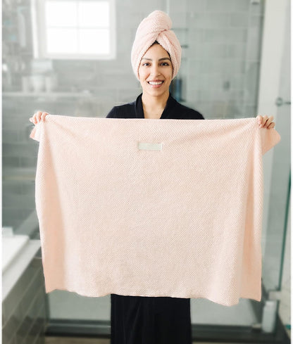 Woman in a dark robe holding up a large light pink textured microfiber hair towel, displayed fully open to show its size. She wears a matching pink hair turban wrap on her head, standing in a bathroom.